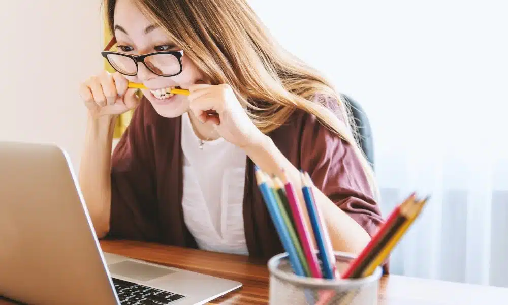 woman biting pencil while sitting on chair in front of computer during daytime